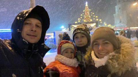 Olesia and her family taking a picture back in Ukraine at Christmas as it snows outside. Pictured from the left to the right is a man in a black coat, a young girl with a red coat, pink scarf and grey hair, a young boy with a black hat and green coat, and Olesia with a hat, black coat and scarf. 