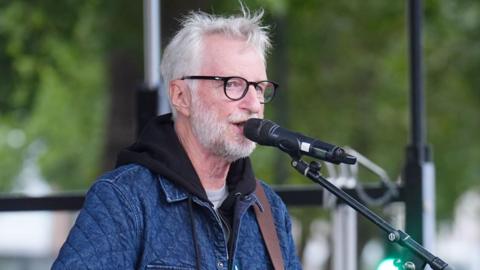 Billy Bragg sings into a microphone as he performs on an outdoor stage. He is wearing a denim-coloured jacked and black-rimmed glasses. He has messy grey hair and a short, full beard.