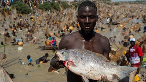 A fisherman poses for a portrait holding a fish he just caught. Blurred in the background dozens of other fishermen can be seen in the water.