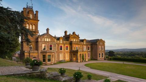 The front aspect of Hestercombe House, a grand, beige-gold stoned building that has a tower to the left. In front of the house is a large driveway with an oval of grassy lawn. Fields and trees extend into the distance.