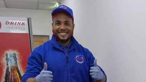 A man with a beard, blue top and blue cap with plastic gloves, doing a thumbs-up with both hands