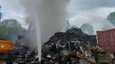 A close up of the fire at the metal recycling centre. Thick, grey smoke is rising from the pile of metal. 