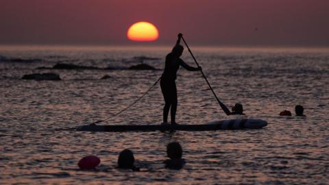 A person standing on a paddle board on the sea is silhouetted against a deep purple sky with the Sun rising on the horizon. Several swimmers are in the water too.