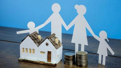 A stock image showing the model of a house next to a pile of coins and a paper cut out of a family.