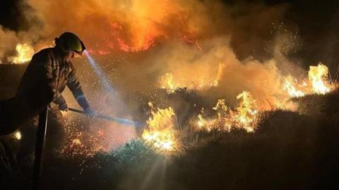 A firefighter with a headtorch using a tool to tackle a gorse fire at night. 