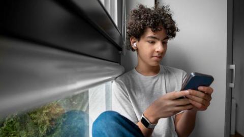 Teenage boy sitting at the window at home using mobile phone.