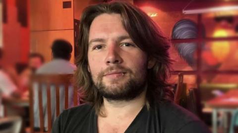 Calum McCombie looking straight at the camera. He has shoulder-length dark hair and a short beard. He has a lip piercing.