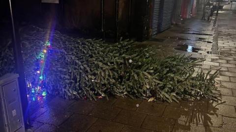 A Christmas tree lying on its side on the street, with only a few lights still working. The street is dark, with scattered puddles. 