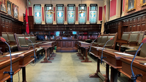 The inside of the House of Keys Chamber, which has green leather clad wooden chairs and benches on either side and a bookcase and stained glass panels behind the speaker's desk.