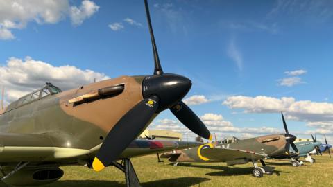 Several warbird planes in a line. They are painted in dark green and brown camouflage with black propellers. They are positioned on grass and behind them is bright blue sky with some white clouds. 