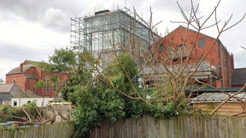 A view of the rear of Jacob's Court in Hereford. Scaffolding covers the tower block and other warehouse buildings are nearby. There is a fence in front of the site, with a tree growing over it.