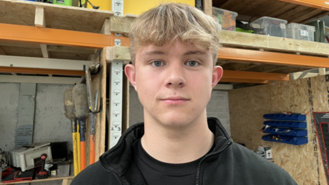 Jack Roberts is looking straight into the camera with a neutral expression. He has short blonde hair with a curly fringe, and is wearing a black t-shirt and black fleece. In the background behind him is the interior of his tool storage unit, where gardening tools including shovels are being stored. 