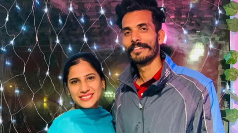 Ravinder Singh wearing a grey jacket with blue stripes standing with his wife wearing a blue scarf around her neck are standing in front of small LED lights 