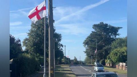 A red and white St George's Flag is tied to a lamppost beside a residential road, and there are two more in the background. There are a couple cars in the frame. There is a blue sky.