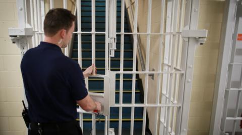 A police warden is closing a white gate inside a prison, with a set stairs behind it.