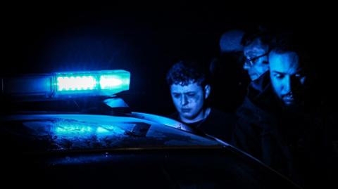 A boy and two men next to a police car, pictured at night with its flashing light on. Their faces are lit up blue and the boy has a bruised face.