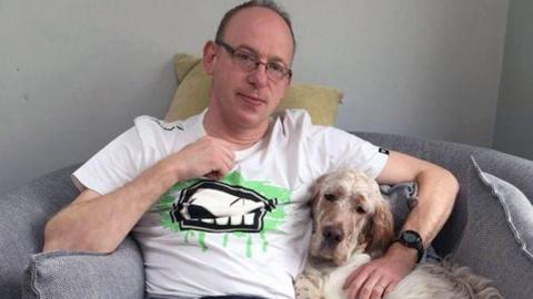 A man in glasses and a white t-shirt sits holding a dog