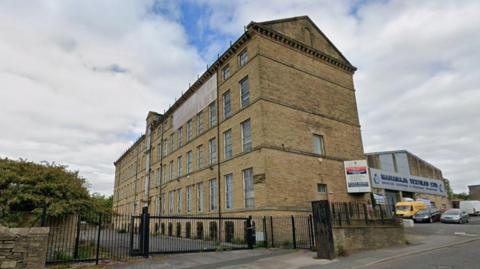 A large, empty mill building behind black gates. A 'To Let' sign covers the top windows of the five-story, light stone building. 