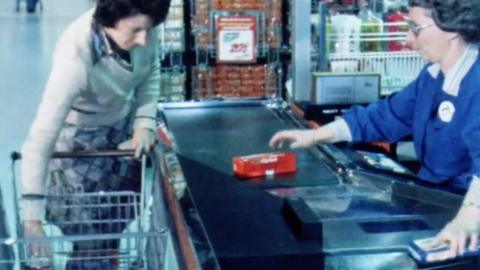 A cashier swipes food products over a laser scanner at a supermarket, while a customer takes more items out of her shopping trolley 