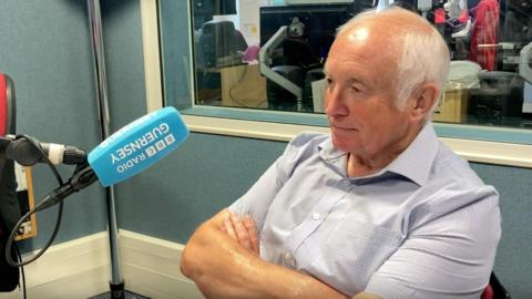 David Harry - a man with white hair, wearing a short-sleeved shirt light blue in the BBC Radio Guernsey studio, arms crossed