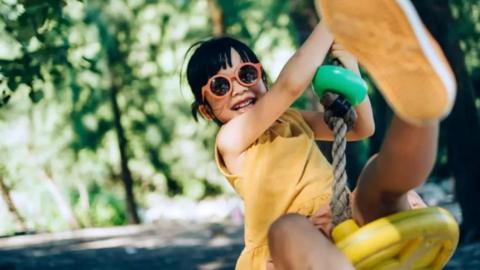 A young girl wearing a yellow dress and sunglasses is on a swing and smiling at the camera