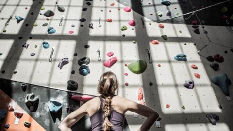 A woman, with her back to the camera, wearing purple vest, looking up at a grey climbing wall, with multi-coloured holds