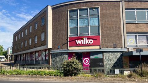 A large brick building with boarded up windows on the ground floor and a red and white sign for Wilko can be seen on a bright sunny day
