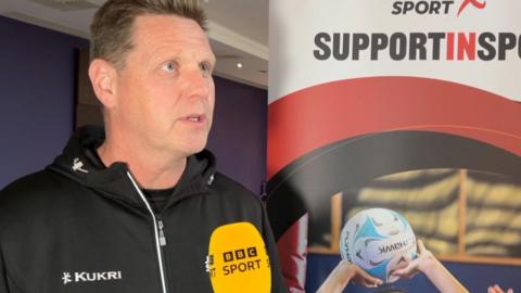 David Kennedy with cropped hair wearing a black tracksuit top is speaking with a yellow BBC Sport microphone in front of him, along with a banner showing two hands holding a ball with a wall in the background painted a shade of purple and mauve with what appears to be a series of ventilation ducts.