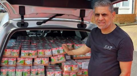 Maqsood Anwar, wearing a black T-shirt, standing by a car that is full of tinned goods. He is looking straight at the camera and has short hair that is slightly greying. 