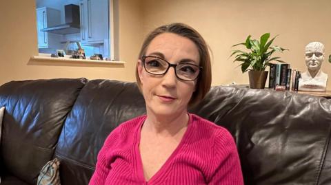 Melissa Finch sits on a brown leather sofa in a living room and poses for a photograph. She has short, cropped brown hair and is wearing dark-rimmed glasses and a fuchsia pink top. A plant can be seen on a table behind her, next to a row of books and a bust of a man's head.