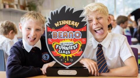 Two pupils in school uniform sat at a desk and sticking their tongues out. There is a trophy in between them with red and black stripes on it, which says "winners Beano Britain's Funniest Class 2026".