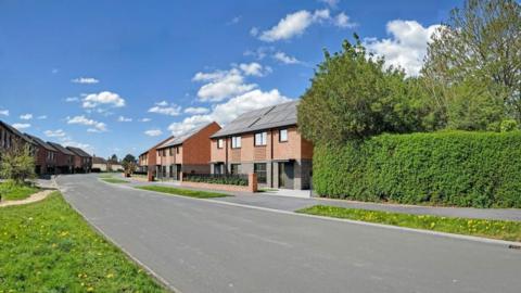 A digitally-generated image of a residential street: brick houses stand among green landscaping.