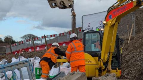 Workers are standing by diggers with a digger arm in the background. They are standing besides another digger.