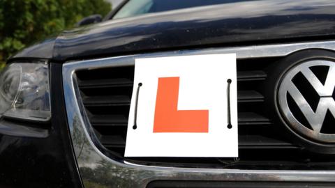 Close up of the grill of a black shiny car with a learner plate tied to it