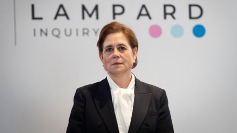 Baroness Kate Lampard wears a black jacket over a white shirt. She stands in front of a white background with the wording "Lampard Inquiry" in black letters, followed by three coloured dots.