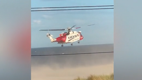A red and white helicopter trying to land on the beach. Sand has been whirled up around the craft and into the sky by the downwind from the blades.