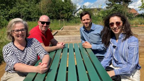 Two men and two women sit at a green picnic bench. In the background are raised beds, wild flowers and trees.