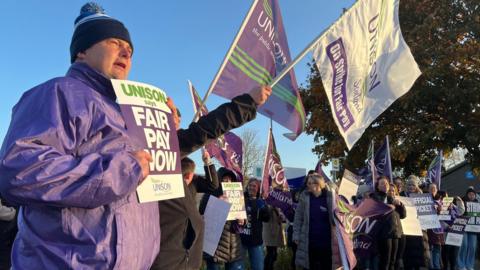 Man in purple jacket and bobble hat holds fair pay now sign as a crowd of picketers behind him wave flagsand banners under a clear blue morning sky.