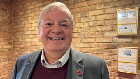 Councillor Alan Amos is smiling at the camera, wearing a grey suit over a maroon jumper. He has short, neatly brushed white hair.