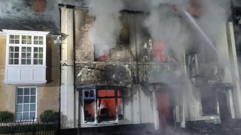 The fire at a property on Northgate Street. Smoke is billowing from the mid-terraced building. Water hoses are being used to put out the fire. The windows of the building have all been smashed and fire can be seen through the windows.