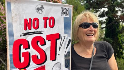 A blonde woman with black sunglasses looking at the camera wearing a black top and with a black strap over her left shoulder. On her right she is holding an AI generated No to GST placard with a Guernsey flag in one corner and a megaphone in the other.