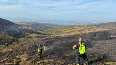 Two firefighters using a hose on a blackened area of moorland