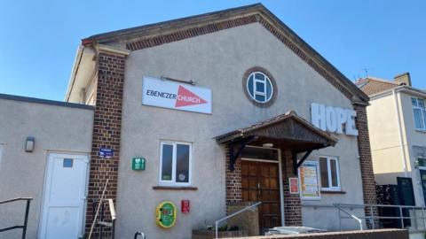 The outside of Ebenezer Church in Horfield, Bristol. The building is beige and has two rectangular windows and a round middle on the front. There is a sign with the church name at the top and a sign reading "HOPE" in capital letters on the side.