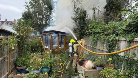 A firefighter is extinguishing the fire at a garden building with a large yellow hose. Smoke is hovering overhead. Smoke can also be seen coming from the roof of a neighbouring shed. The garden is full of pots of flowers and greenery. It is an overcast day.