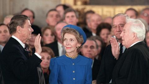 Ronald Reagan holds up his right hand at his swearing in as US President next to his wife Nancy with a crowd around him.