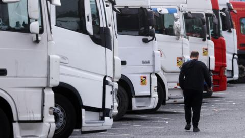 Man walking past a line of white lorries with his back to the camera. He is wearing all black.