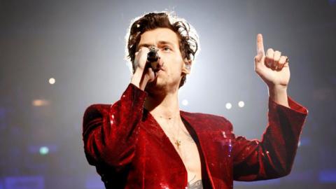Harry Styles sings into a microphone while performing at the Brit awards in 2023. He has a red, sequinned jacket on and is pointing one finger up towards the ceiling.