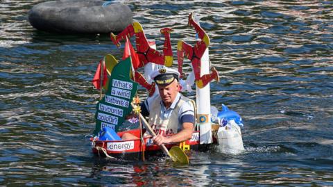 A man in a decorated tin bath rows through the water. It has two large triskelions attached to it, and a map of the Isle of Man.