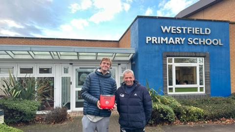 Johannus Monday with head coach Paul Holtby holding a gifted defibrillator from his former primary school