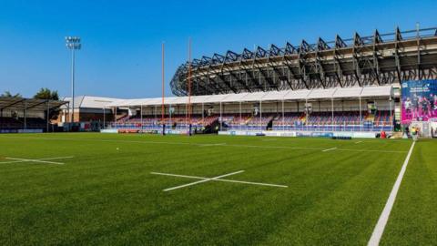A sunny, empty rugby stadium with a green pitch, goalposts, and seating stands under a bright blue sky.